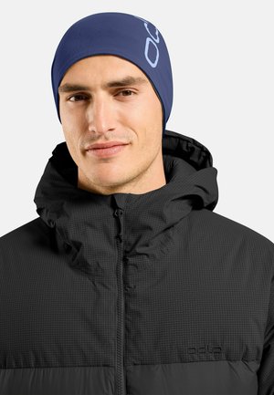 Man wearing a navy blue beanie and zipped black quilted jacket with high collar, facing forward against a plain background.