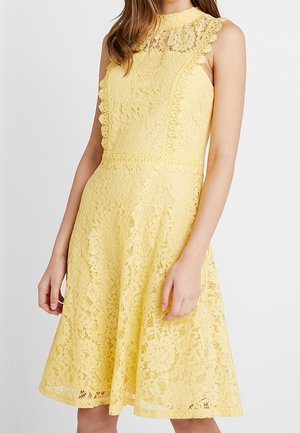 Geel kantjurk met een hoge halslijn, geschulpte randen en een uitlopende rok. Intricate bloemenpatronen door de stof heen.