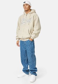 Felpa beige in pile con zip e logo, jeans oversize blu a fantasia e sneakers bianche. Il modello indossa un berretto bianco a coste.