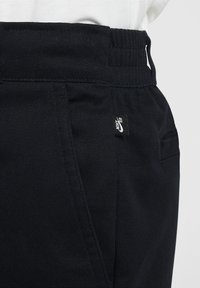 Čierne nohavice s rebrovaným pásom, bočnými vreckami a malou nášivkou Nike SB. Tkanina má hladkú textúru. Jednoduchý, moderný dizajn.