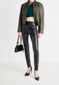 Kvinna i mörkgrön kort jacka, grön croptop, svarta läderbyxor, svarta högklackade skor, som håller en liten svart handväska mot en vit bakgrund.