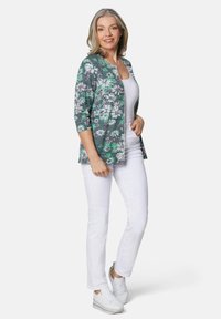 Blumenmuster-Jacke in Grün, Pink und Weiß mit Dreiviertelärmeln, kombiniert mit weißen Hosen und weißen Sneakers.