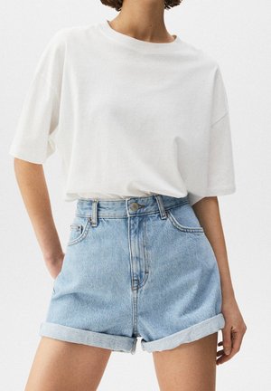 Personne portant une chemise blanche ample à manches courtes rentrée dans un short en jean bleu clair avec ourlet retroussé, debout les mains partiellement visibles.