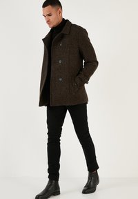 Braune, strukturierte Jacke mit Spitzkragen, zwei großen Knöpfen, Seitentaschen und taillierter Silhouette, kombiniert mit schwarzen, schmalen Hosen und Ankle Boots.