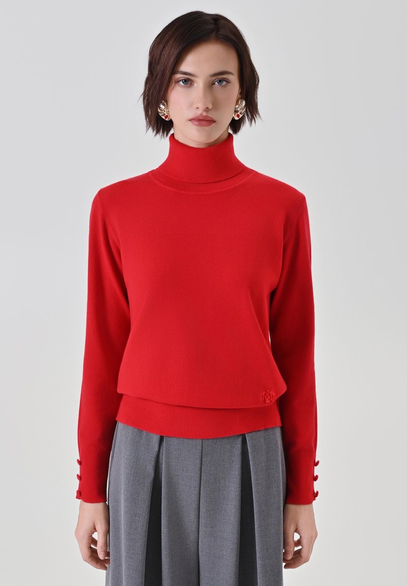 Roter Rollkragenpullover mit langen Ärmeln, geripptem Muster und Knopfakzenten an den Manschetten. Kombiniert mit grauen, gefalteten Hosen.