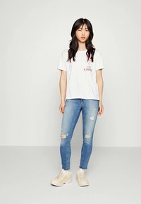 White cotton t-shirt with printed text, paired with light blue ripped skinny jeans and beige sneakers. Straight hem and relaxed fit.