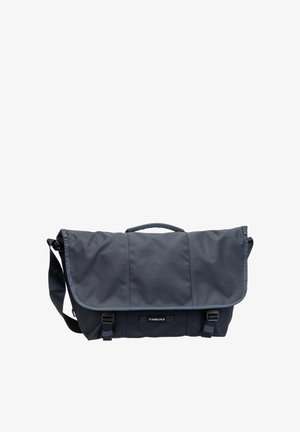 Dunkelblaue Messenger-Tasche mit verstellbarem Schultergurt, oberem Griff und zwei Schnallenverschlüssen, vorne mit der Aufschrift „TIMBUK2“.