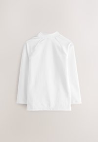 Vit långärmad skjorta i mjuk bomull med hög krage, raglanärmar och förstärkta sömmar längs fåll och ärmslut.