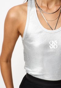 Zilveren mouwloze crop top van ribbelmateriaal met een gestructureerde afwerking en een wit logo; gelaagde zilveren kettingen als extra detail.