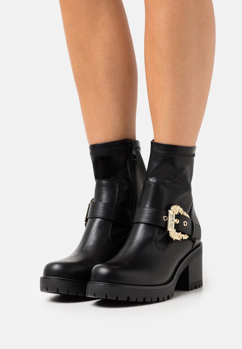 Botas de tobillo negras de cuero liso, con una hebilla dorada decorativa, suela de goma y un tacón grueso para mayor estabilidad.