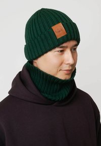 Zielona dzianinowa czapka z prążkowaną fakturą i brązową skórzaną naszywką. W zestawie z dopasowanym zielonym prążkowanym otulaczem na szyję.