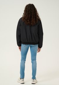 Giacca bomber nera in nylon con polsini e orlo elasticizzati, indossata sopra jeans skinny blu chiaro e sneakers bianche, vista da dietro.