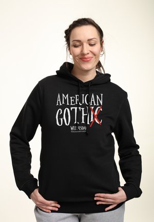 Schwarzer Hoodie mit durchgestrichenem "AMERICAN GOTH", ausgestattet mit einer Kordelzugkapuze, langen Ärmeln und einem lockeren Schnitt. Weicher Stoff mit minimaler Textur.
