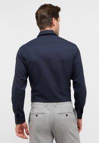 Uomo che indossa una camicia a maniche lunghe blu navy aderente e pantaloni eleganti grigi chiaro, mostrato da dietro su uno sfondo neutro.