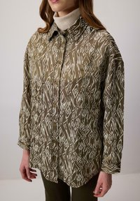 Camicia verde a fantasia con bottoni sul davanti, caratterizzata da forme a diamante bianche. Tessuto leggero, vestibilità rilassata, maniche lunghe e taschino sul petto.