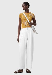 Geel bloemmotief kortmouwen top gecombineerd met hooggetailleerde witte geplisseerde broek en platte sandalen met zilveren accenten. Eenvoudige grijze achtergrond.