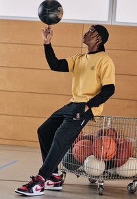 Basketbola spēlētājs, tērpies dzeltenā īsā piedurkņu krekla virs melnā garā piedurkņu krekla, melnās biksēs un sarkani/melnos sporta apavos, līdzsvaro basketbola bumbu.