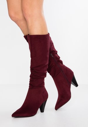 Mujer con botas de ante burdeos, de caña alta y caída, con cremallera lateral y tacones en forma de cono, sobre un fondo blanco.