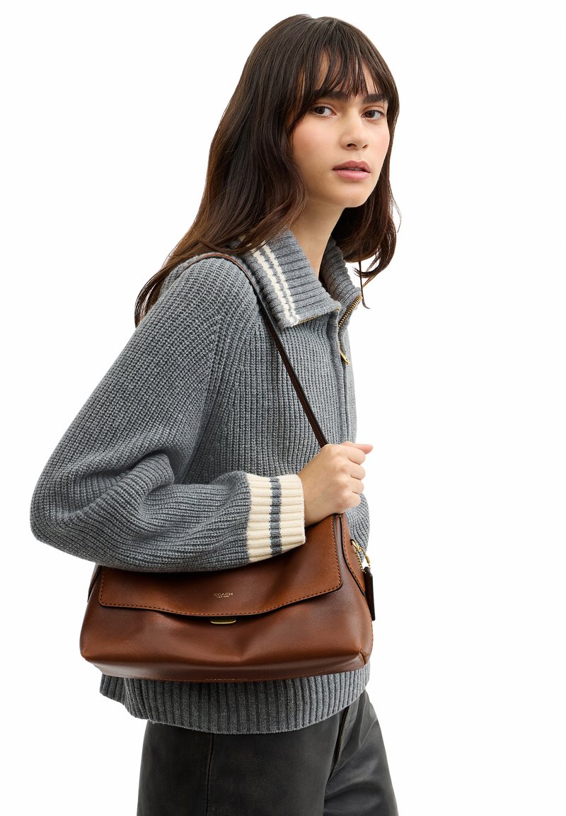 Femme portant un pull en tricot gris tient un sac à bandoulière en cuir marron avec une boucle dorée, debout devant un fond blanc.