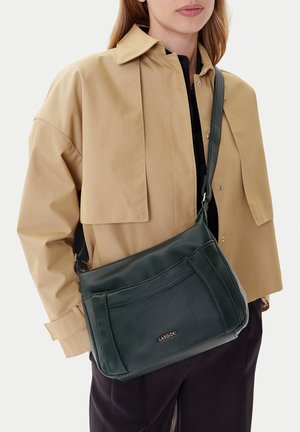Woman wearing beige jacket and black pants carrying dark green Lasocki leather shoulder bag across body.