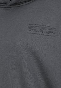 Grauer Hoodie mit weicher Textur. Auf der Vorderseite befinden sich schwarze, aufgedruckte Texte mit Phrasen über Glück und Selbstfürsorge.