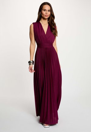 Kvinde med langt brunt hår iført ærmeløs dyb lilla plisseret maxikjole og sølvarmbånd, stående mod en ensfarvet lys baggrund.