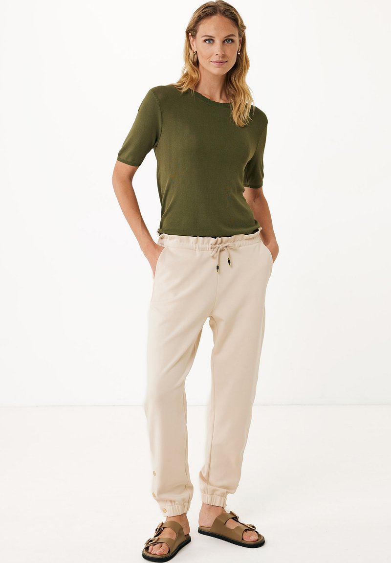 Olijfgroen gebreide top, lichtbeige joggers met elastische boorden en bruine sandalen. Bevat een trekkoord in de taille en knoopdetails op de broek.