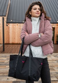 Black fabric tote bag with two black straps, featuring a pink logo. Held by a person in a light pink puffer jacket and a white knit sweater.