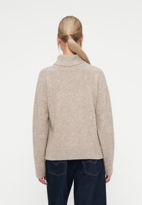 Beige kötött pulóver magas gallérral, süllyesztett vállvarrásokkal, bordás mandzsettákkal és alsó szegéllyel, sötétkék, magas derekú farmernaci mellett viselve.