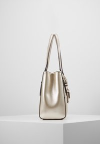 Metallinen kultainen käsilaukku, jossa on sileä pinta, jämäkkä muoto ja kaksi pitkää kahvaa. Varustettu hillityillä saumoilla ja pienellä lukkoyksityiskohdalla.