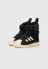Botas aislantes negras con puños elásticos, puntera de goma blanca, tres rayas blancas y detalle de correa lateral. Parte superior de tela texturizada.