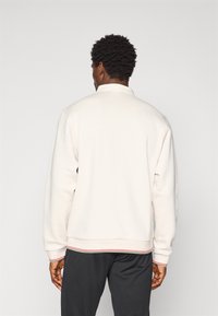Lys beige sweatshirt med høj krave, ribbet manchetter og en rød og grøn stribet accent ved sømmen. Blød, tekstureret stof.