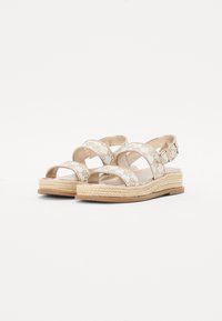Kile-sandaler med kryssende stropper og spenne. Laget av lyse beige materiale med en vevd jute-såle og subtilt preget mønster.