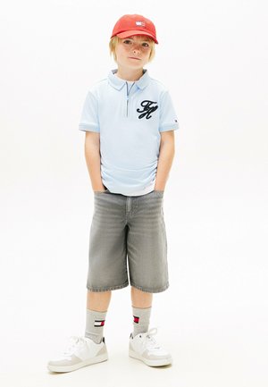 Junger Junge mit blondem Haar, trägt eine rote Mütze, hellblaues Poloshirt, graue knielange Shorts, graue Socken und weiße Turnschuhe, steht mit den Händen in den Taschen.