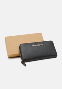 Črn usnjen denarnica z zadrgo z teksturirano površino, z zlatim natisnjenim logotipom "Valentino" in zlatim potegom za zadrgo. Pritrjena je v rjavem kartonskem škatli.