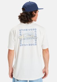Hvid kortærmet t-shirt med en blå grafik p�å ryggen, der viser et bølgedesign og forskellige mønstre. Stoffet ser blødt og let ud.