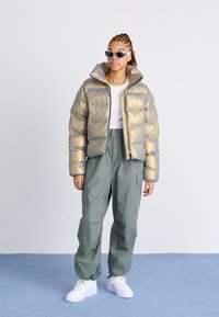 Giacca grigia e oro imbottita con colletto alto, abbinata a pantaloni cargo verde chiaro e sneakers bianche. Il modello indossa occhiali da sole scuri.