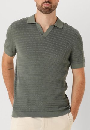 Mann trägt ein kurzärmeliges, strukturiertes Poloshirt in Olivgrün mit V-Ausschnitt-Kragen, kombiniert mit hellbeigen Hosen.