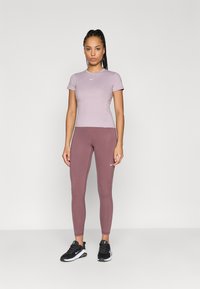 Ανοιχτό μωβ αθλητικό μπλουζάκι με κοντά μανίκια και στρογγυλή λαιμόκοψη, σε συνδυασμό με εφαρμοστά σκούρα μοβ κολάν και μαύρα αθλητικά παπούτσια με λευκές λεπτομέρειες.