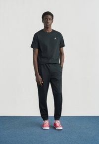 Μαύρο βαμβακερό μπλουζάκι και φόρμα με ελαστική μέση, σε συνδυασμό με κόκκινα αθλητικά παπούτσια με λευκή σόλα και κορδόνια, σε φόντο μπλε χαλί.