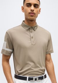 Άνδρας που φοράει μπεζ πόλο με γιακά που κουμπώνει και λεπτομέρεια με ρίγες στο μανίκι, συνδυασμένο με λευκό παντελόνι και μαύρη ζώνη.