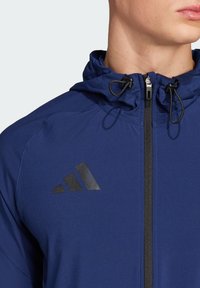 Marineblauwe hoodie met een volledige rits, zwarte accenten en een reflecterende Adidas-logo op de borst, met een gestructureerde stof.