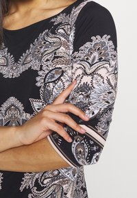 Černý top s jemnými květinovými a listovými vzory v béžové, šedé a světle růžové barvě. Má tříčtvrteční rukávy a kulatý výstřih.