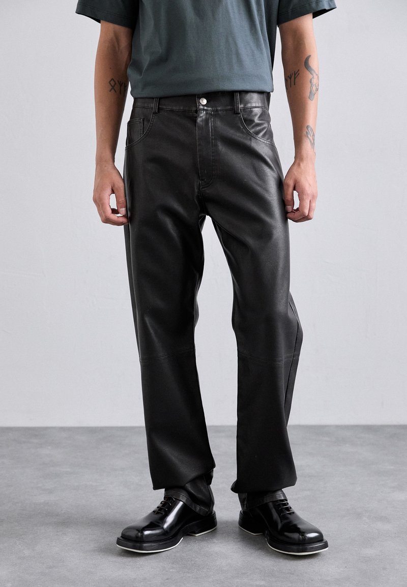 Pantalones de cuero negros con un ajuste relajado, que presentan costuras visibles, bolsillos y un cierre de botón. Acompañados de zapatos de cuero negros.