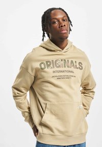 Beige hoodie with a drawstring, front pocket, and printed text "ORIGINALS INTERNATIONAL" in contrasting colors. Cotton blend material, relaxed fit.