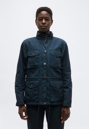 Persoană care stă în picioare cu fața înainte, purtând o jachetă albastru închis cu patru buzunare cu clapetă fixate cu nasturi și un guler înalt.