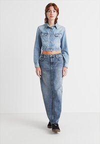 Giacca di jeans blu chiaro con design cropped e due tasche sul petto, abbinata a una gonna di jeans blu chiaro a vita alta e scarpe nere.