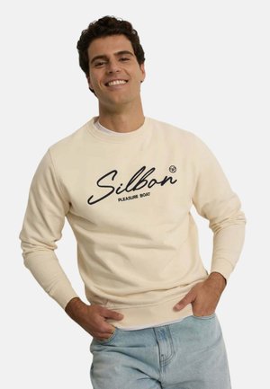 Hombre sonriente que lleva una sudadera crema con el texto "Silbon Pleasure Boat", de pie con las manos en los bolsillos de unos vaqueros azul claro.