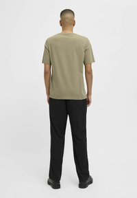 Camiseta de algodón verde oliva con mangas cortas y cuello redondo, combinada con pantalones negros de corte holgado y zapatos oscuros, vista desde la espalda.