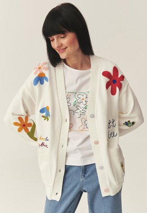 Žena v bílém kardiganu s barevnými květinovými nášivkami a vyšívaným nápisem, v kombinaci s tričkem s květinovým potiskem a světle modrými džínami, usmívající se.